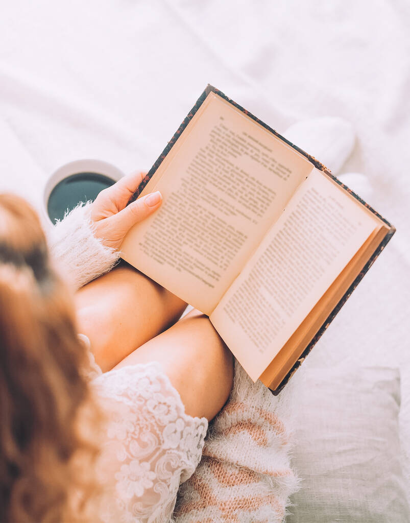 Młoda kobieta na łóżku ze starą książką i filiżanką kawy cieszy się pobytem. - Zdjęcie, obraz