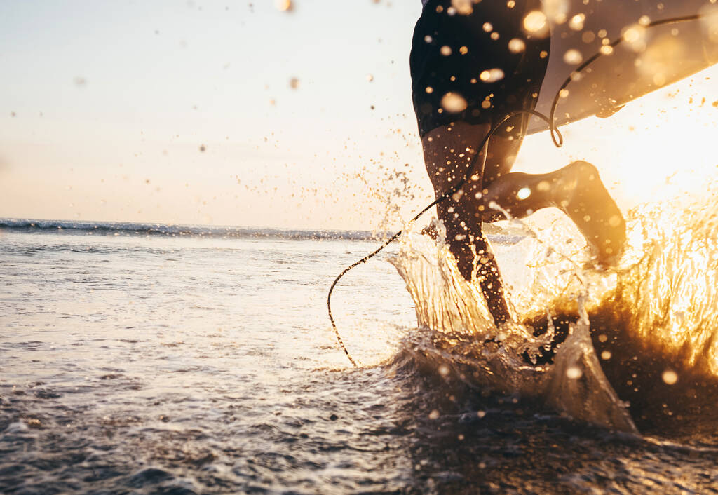 Hombre surfista correr en el océano con tabla de surf. Imagen de primer plano salpicaduras de agua y piernas, luz del atardecer
 - Foto, Imagen