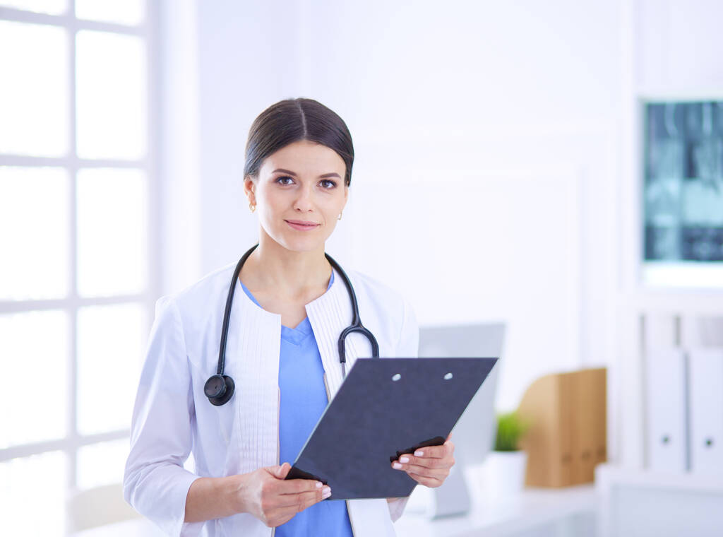 Молодая улыбающаяся женщина-врач со стетоскопом держит папку в кабинете врача - Фото, изображение