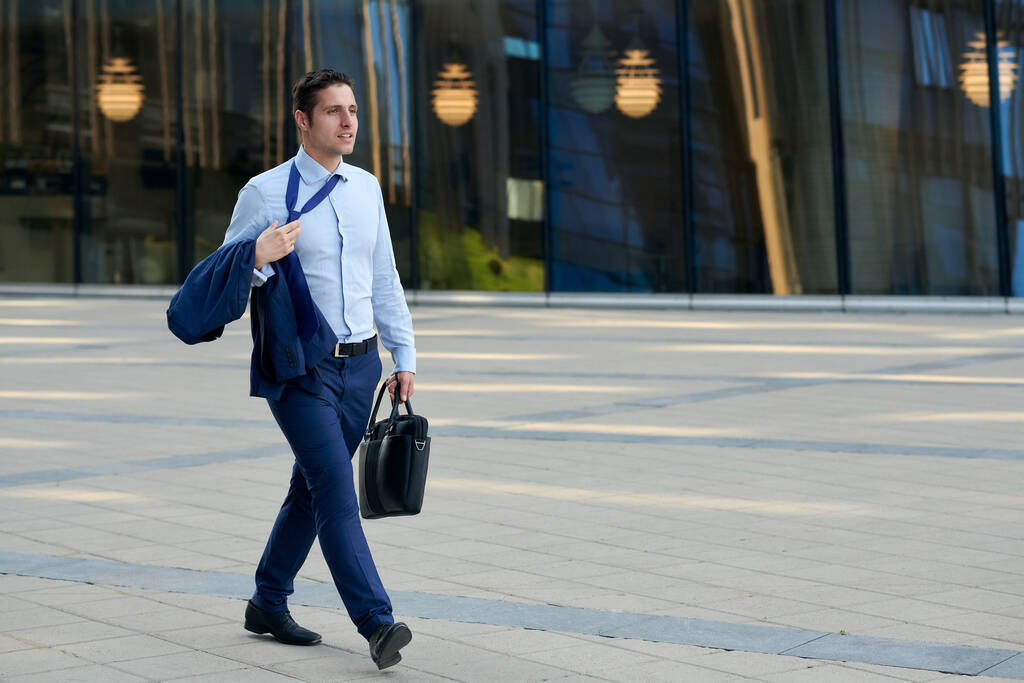 Κουρασμένος νεαρός επιχειρηματίας με υπόθεση πηγαίνει μετά την εργασία σε ζεστό καιρό   - Φωτογραφία, εικόνα