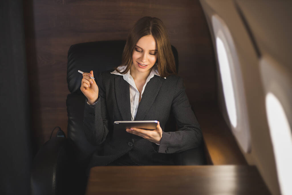 Hermosa exitosa joven empresaria de cabello castaño vistiendo traje negro, usando tableta, sonriendo mientras está sentada en la silla de su avión privado de negocios. Volar en primera clase
. - Foto, imagen