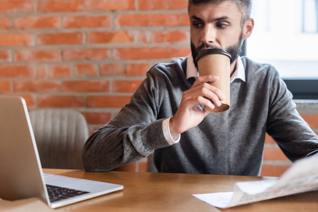 избирательный фокус привлекательного бизнесмена, смотрящего на ноутбук, держа газету и выпивая кофе, чтобы пойти
 - Фото, изображение