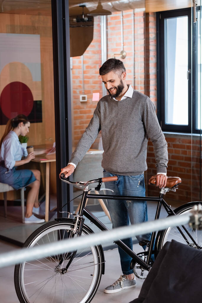 избирательный фокус счастливого бизнесмена, стоящего на велосипеде рядом с бизнес-леди, сидящей в офисе
 - Фото, изображение