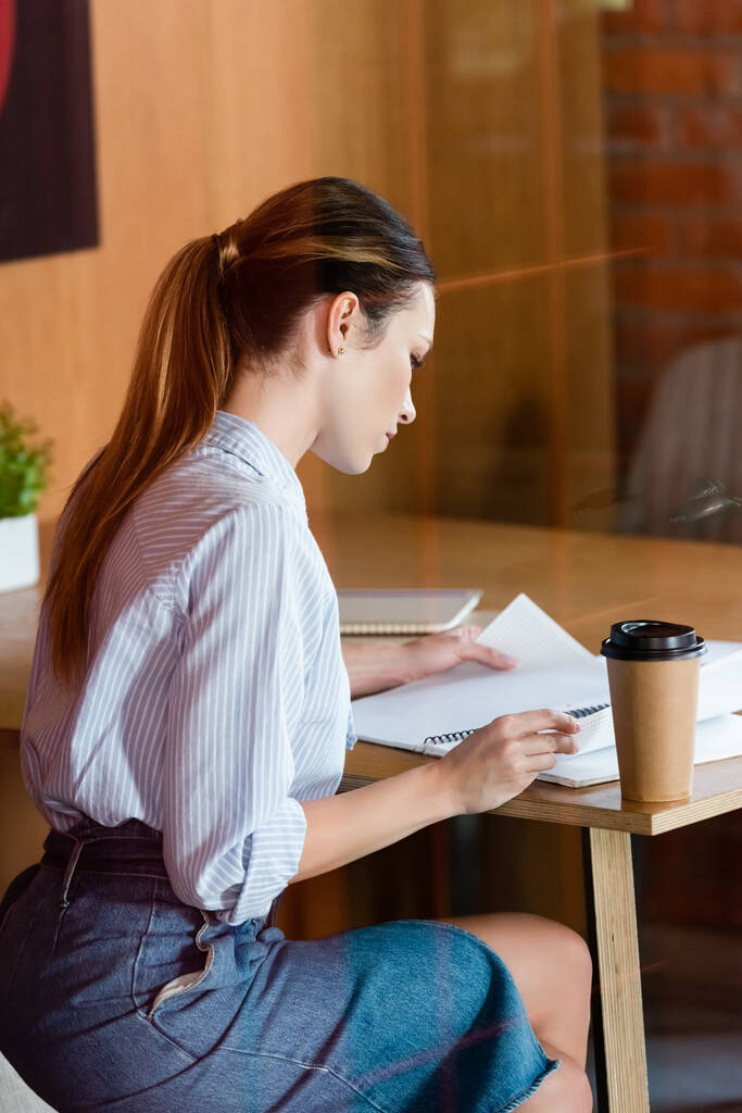 femme d'affaires assise près d'une tasse en papier avec du café à emporter tout en regardant un carnet  - Photo, image
