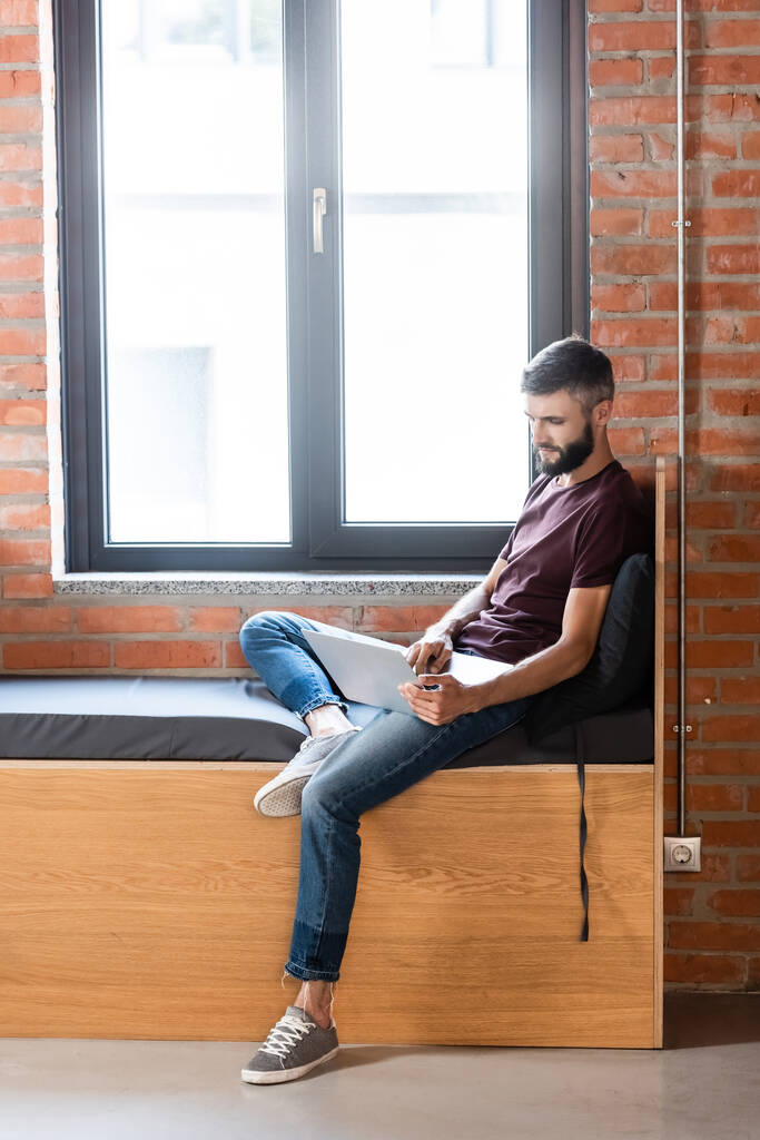 pohledný podnikatel pomocí notebooku, zatímco sedí na lavičce v moderní kanceláři - Fotografie, Obrázek