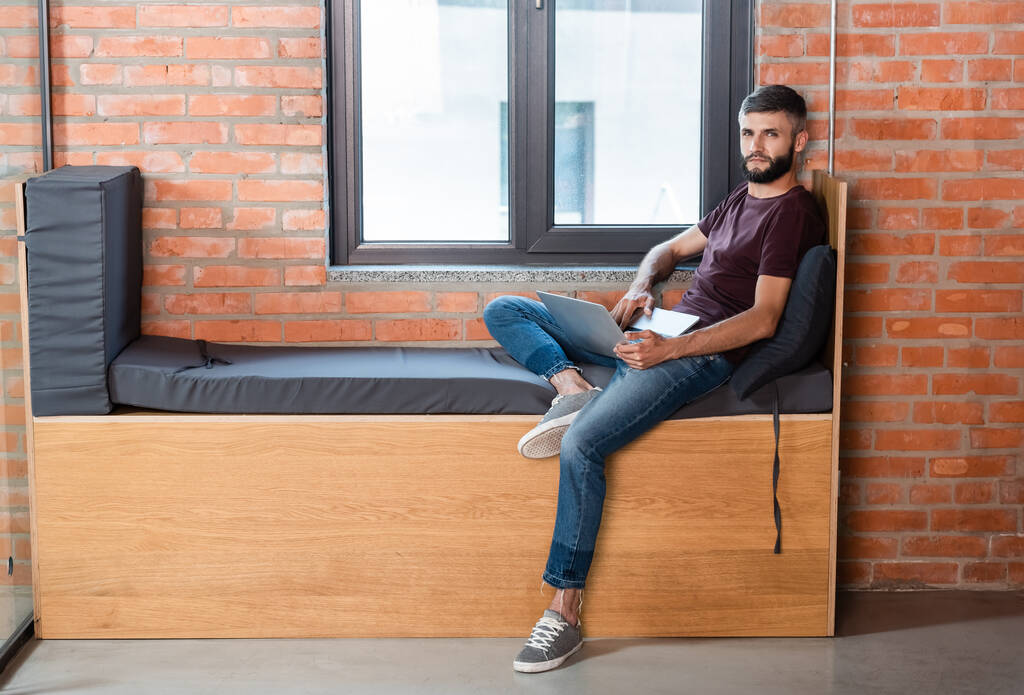 szakállas üzletember ül laptoppal ablak padon a modern irodában - Fotó, kép