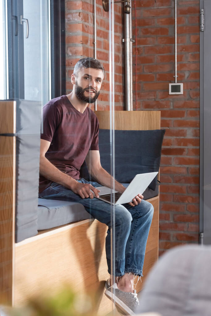 selektiver Fokus eines bärtigen Geschäftsmannes, der auf einer Fensterbank sitzt und einen modernen Laptop hält, während er in die Kamera blickt - Foto, Bild