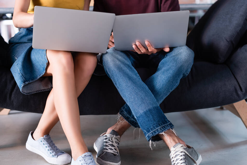 abgeschnittene Ansicht von Geschäftsmann und Geschäftsfrau, die Laptops benutzen, während sie auf dem Sofa sitzen - Foto, Bild