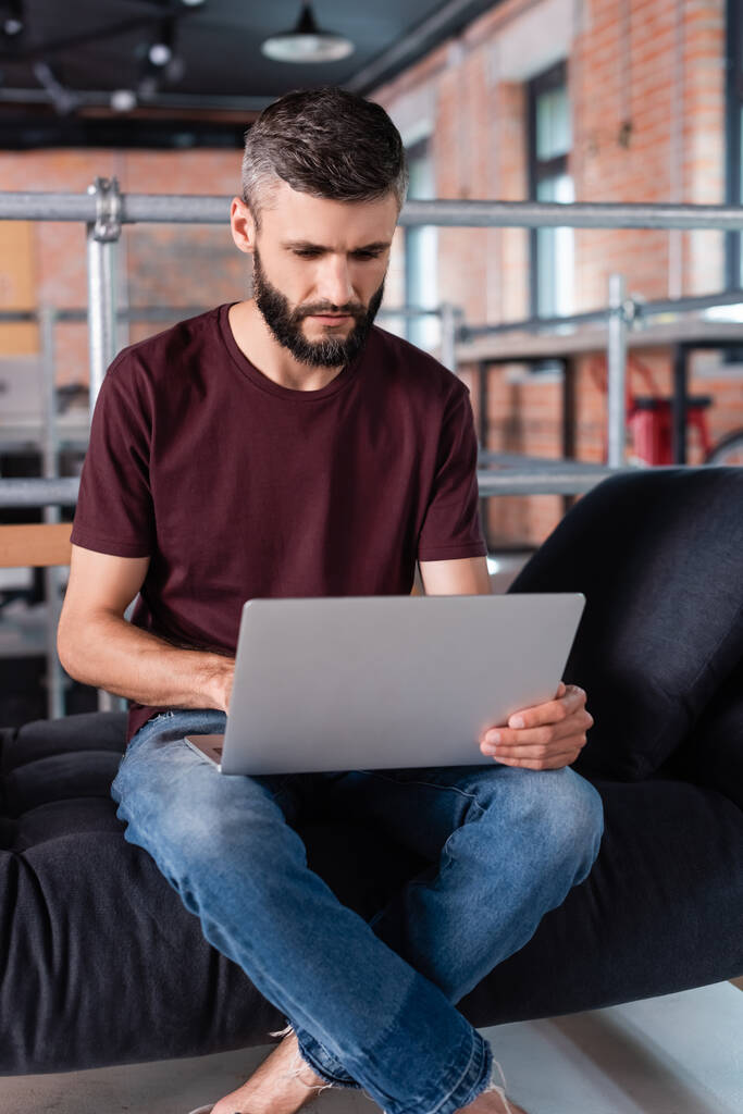 pohledný podnikatel se zkříženýma nohama sedí na pohovce a pomocí notebooku v kanceláři - Fotografie, Obrázek