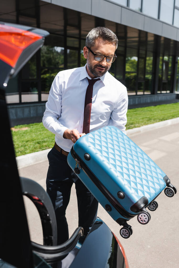 Selective focus of handsome businessman holding suitcase near car with open trunk on urban street  - Photo, Image