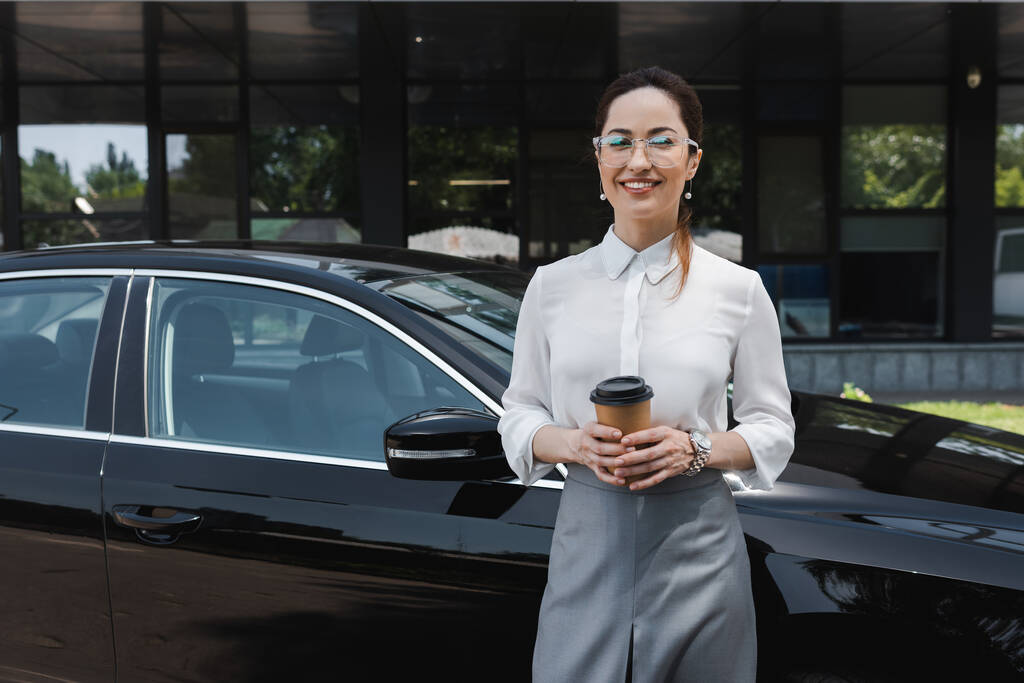 Bella donna d'affari sorridente alla macchina fotografica, mentre tenendo tazza di carta vicino auto sulla strada urbana  - Foto, immagini