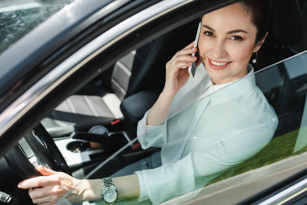Selektiver Fokus der Geschäftsfrau, die in die Kamera lächelt und auf dem Fahrersitz im Auto mit dem Smartphone spricht  - Foto, Bild