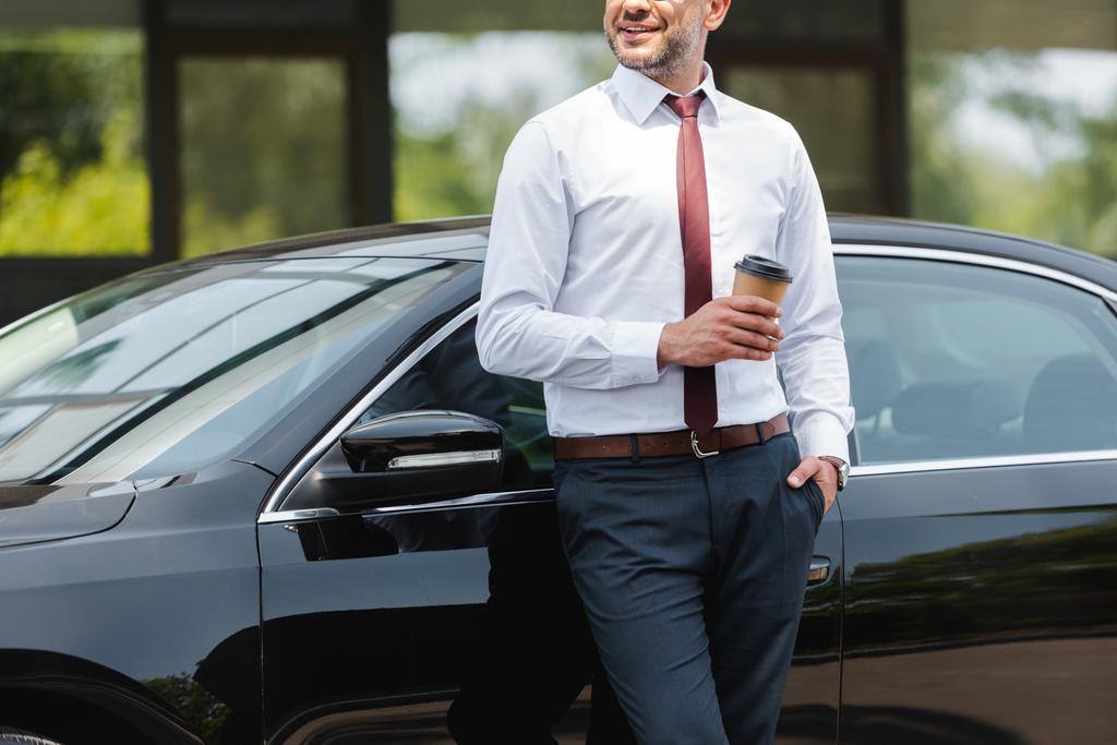 Vista ritagliata dell'uomo d'affari sorridente che tiene la tazza di carta vicino all'auto sulla strada urbana  - Foto, immagini