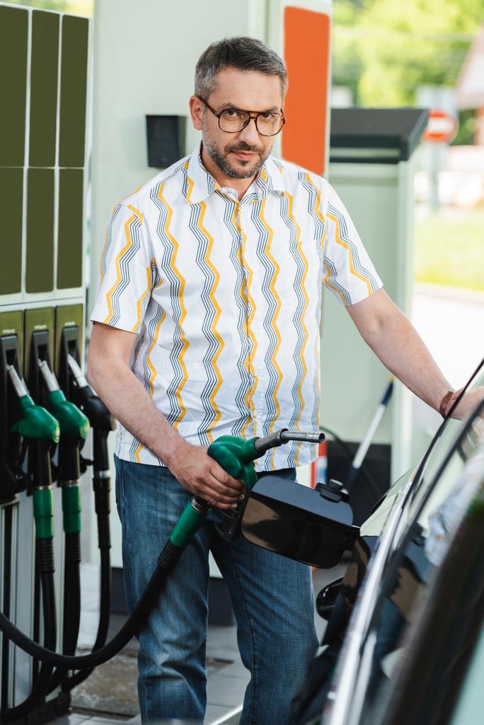 Szelektív fókusz jóképű férfi nézi a kamerát, miközben tartja üzemanyag fúvóka autó közelében autó töltőállomás - Fotó, kép