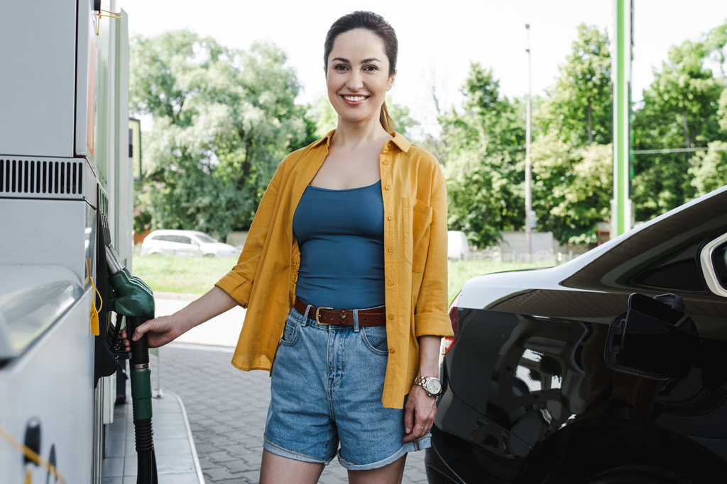 ガソリンスタンドで車の近くに燃料ノズルを保持する美しい笑顔の女性の選択的な焦点  - 写真・画像