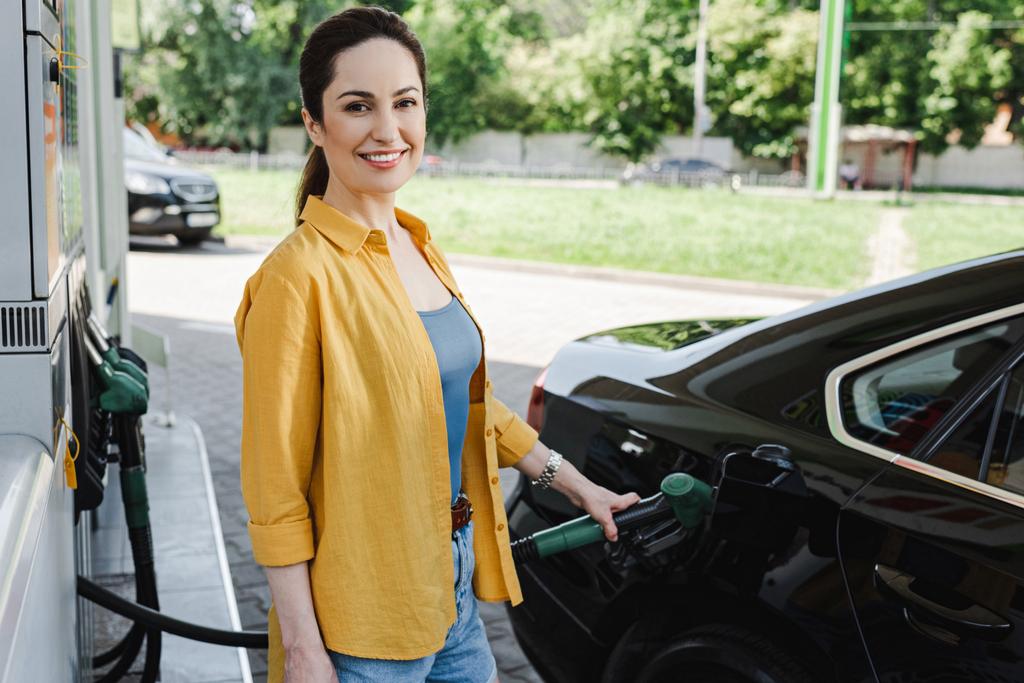 A kamerába mosolygó nő szelektív fókusza tankolás közben a benzinkúton  - Fotó, kép