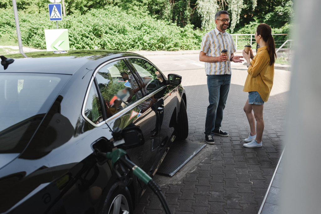 Selective focus of smiling man holding paper cup near wife while fueling car on gas station  - Photo, Image