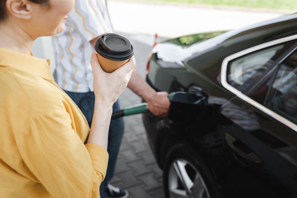 Ausgeschnittene Ansicht einer Frau, die Kaffee to go hält, während ihr Mann Auto an Tankstelle tankt  - Foto, Bild