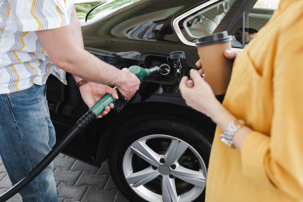 Widok człowieka trzymającego dyszę paliwową w pobliżu zbiornika paliwa samochodu w pobliżu kobiety z papierową filiżanką na stacji benzynowej  - Zdjęcie, obraz