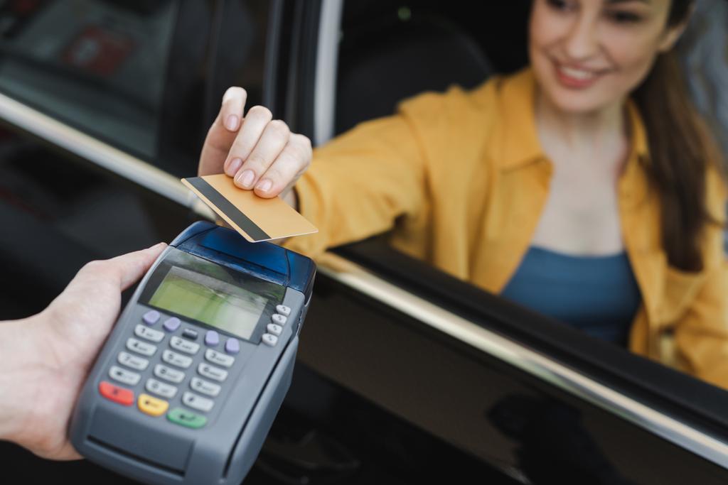 Szelektív fókusz mosolygó nő kezében hitelkártya, miközben ül az autóban, közel a munkavállaló a benzinkút gazdaság fizetési terminál - Fotó, kép