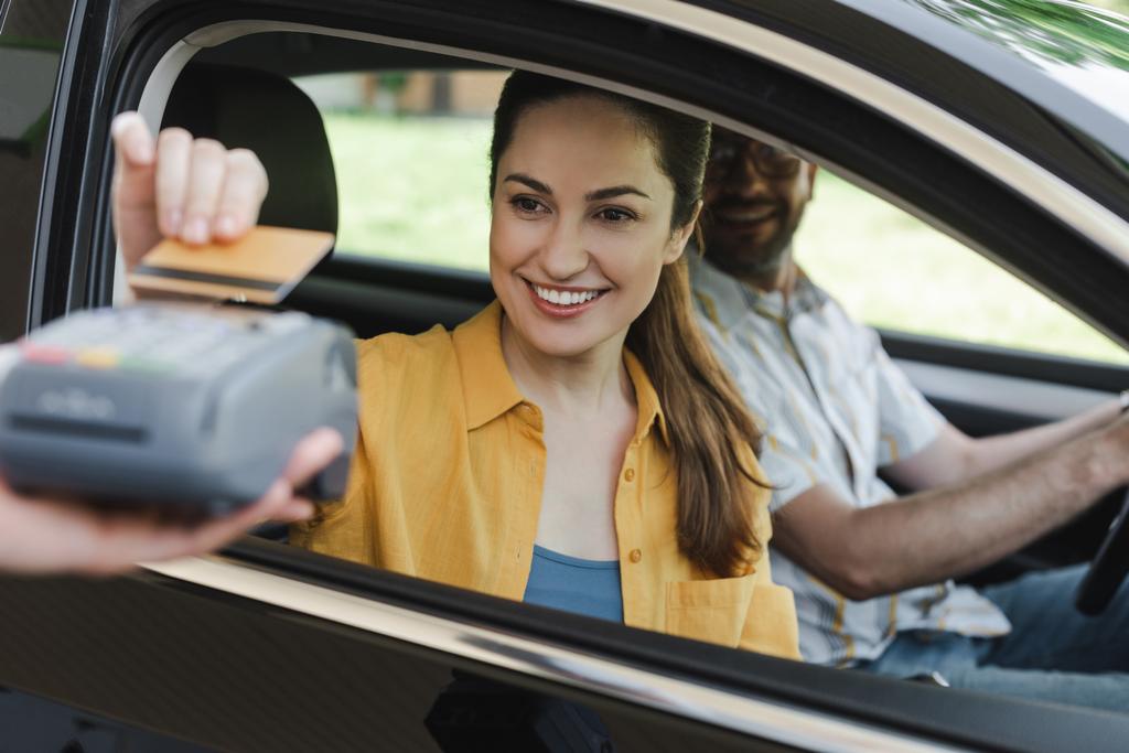 Szelektív fókusz a mosolygó nő fizet hitelkártyával közeli férj az autóban és a munkavállaló a benzinkút gazdaság fizetési terminál - Fotó, kép