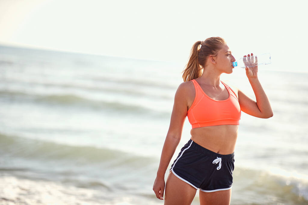 молода біла біла жінка п'є воду з пляшки на пляжі, роблячи паузу від вправ
 - Фото, зображення