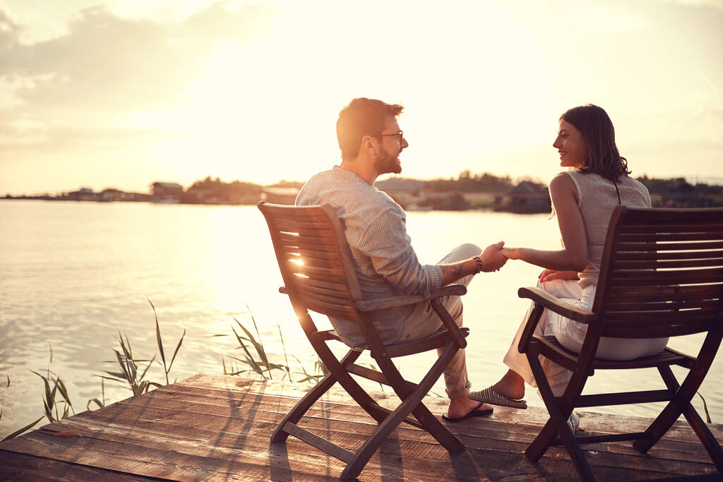 romantyczna, kaukaska para, siedząca na krzesłach nad rzeką, trzymająca się za ręce, rozmawiająca, obserwująca się nawzajem. przestrzeń kopiowania - Zdjęcie, obraz