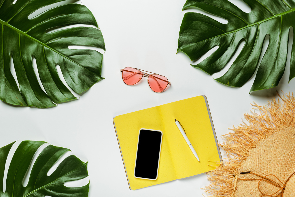 widok z góry zielone liście palmy, słomy kapelusz, okulary przeciwsłoneczne i żółty notatnik ze smartfonem na białym tle - Zdjęcie, obraz