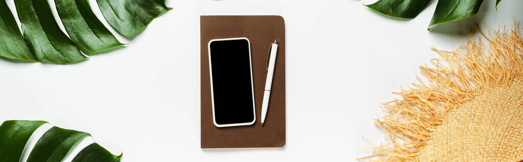 πάνω άποψη του πράσινου φύλλα φοίνικα, ψάθινο καπέλο και σημειωματάριο με smartphone σε λευκό φόντο, πανοραμική λήψη - Φωτογραφία, εικόνα
