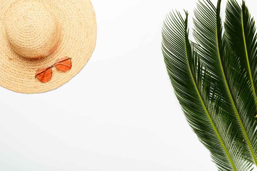 vrchní pohled na zelené palmové listy v blízkosti slaměný klobouk a sluneční brýle na bílém pozadí - Fotografie, Obrázek