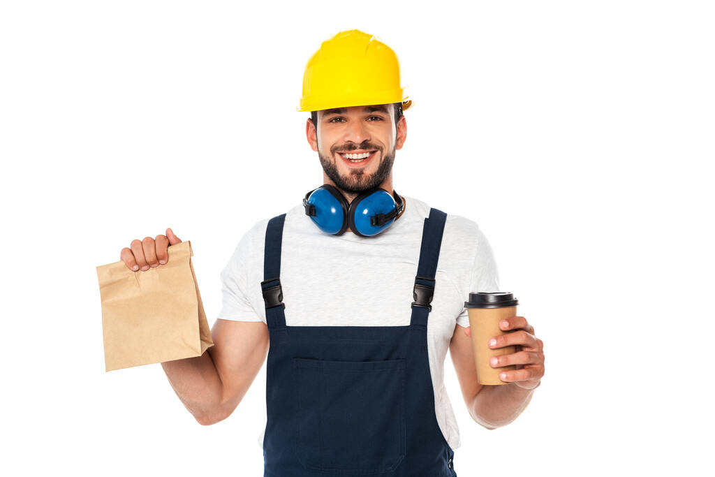 Schöner Arbeiter, der Coffee to go und Papiertüte in der Hand hält und isoliert auf Weiß in die Kamera lächelt - Foto, Bild
