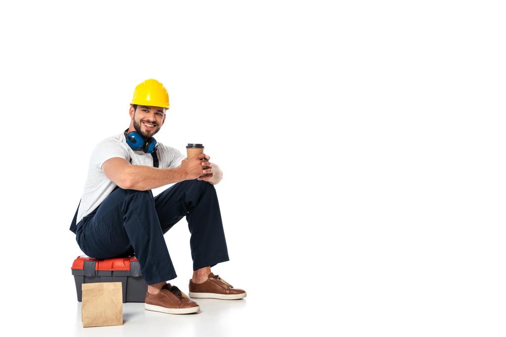 Reparador sonriente en uniforme sosteniendo café para llevar mientras está sentado en la caja de herramientas cerca de la bolsa de papel sobre fondo blanco
 - Foto, imagen