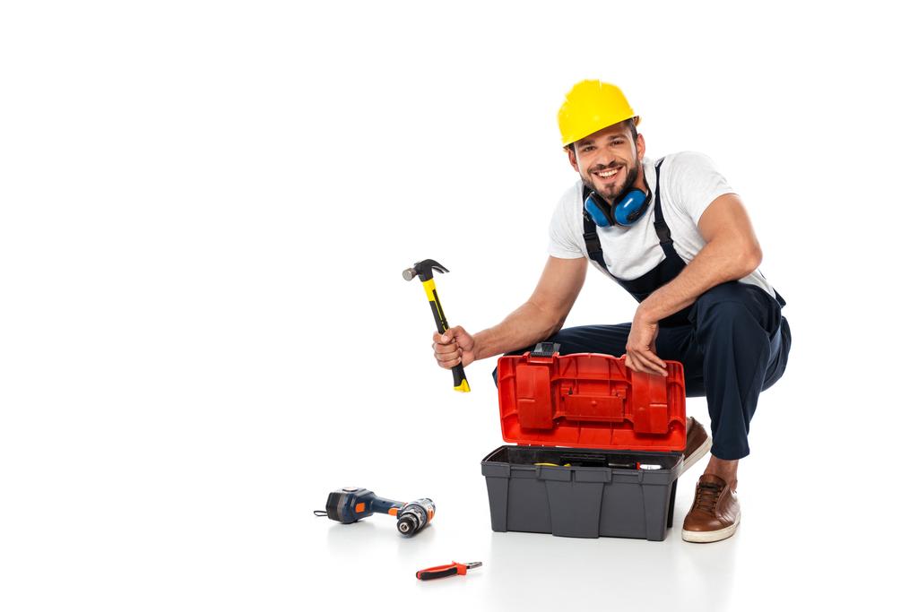 Glimlachende werkman in uniforme houthamer in de buurt van gereedschapskist en gereedschappen op witte achtergrond - Foto, afbeelding