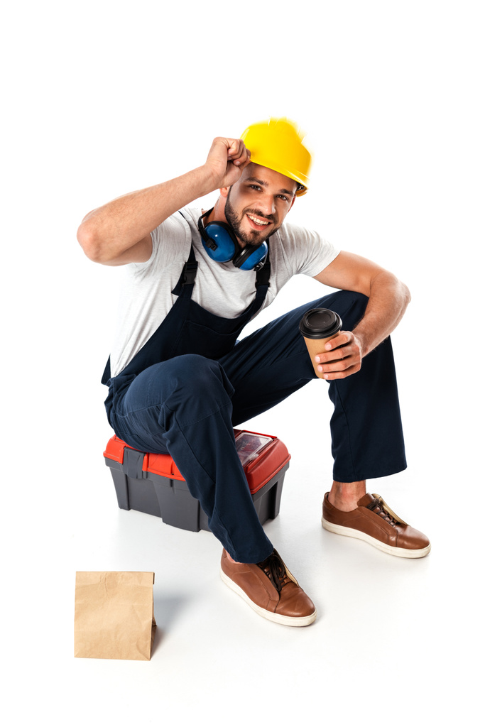 Trabalhador sorridente em uniforme e chapéu duro segurando café para ir enquanto sentado na caixa de ferramentas perto de saco de papel no fundo branco
 - Foto, Imagem