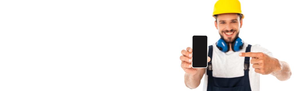 Panoramische oogst van lachende klusjesman wijzend met de vinger naar smartphone met blanco scherm geïsoleerd op wit - Foto, afbeelding