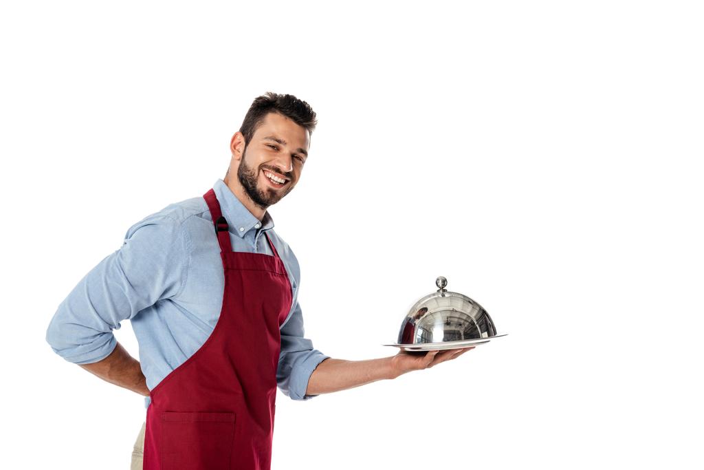 Σερβιτόρος σε ποδιά κρατώντας μεταλλικό κάλυμμα πιάτου και δίσκο, ενώ χαμογελά στην κάμερα απομονώνονται σε λευκό - Φωτογραφία, εικόνα