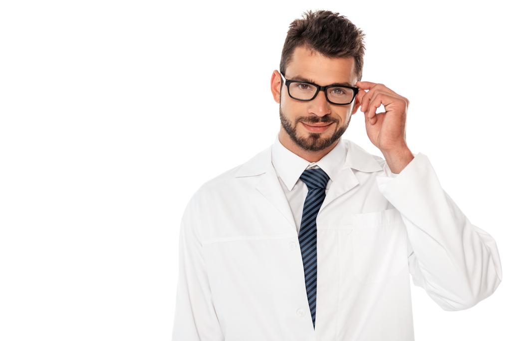 Usmívající se vousatý lékař drží brýle a usmívá se na kameru izolované na bílém - Fotografie, Obrázek