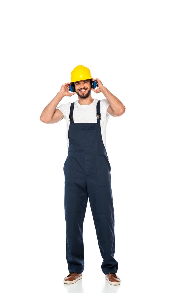 Glimlachende werkman in hardhat en oorbeschermers kijkend naar camera op witte achtergrond - Foto, afbeelding
