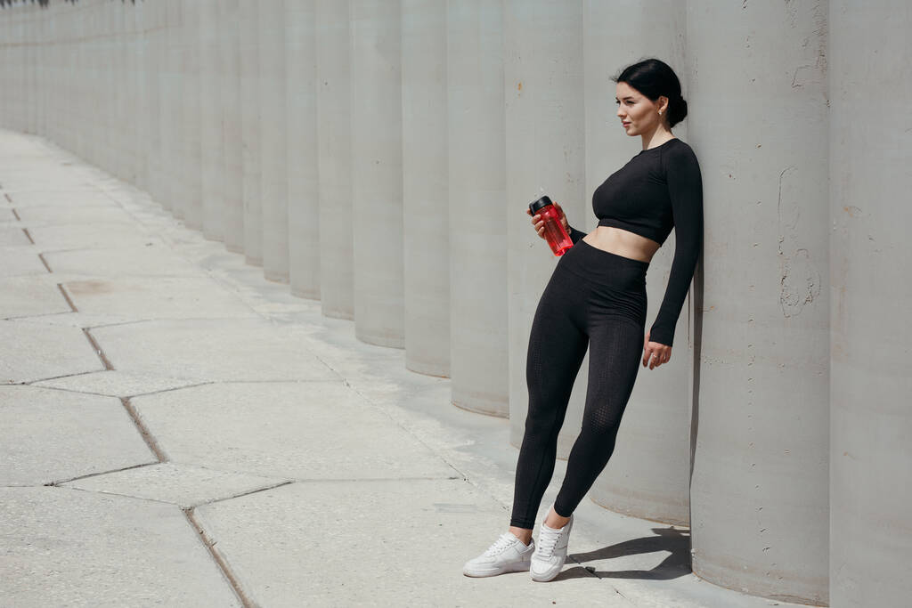 Девушка в облегающем черном спортивном костюме прислоняется к бетонной стене и держит в руке бутылку воды на улице.
. - Фото, изображение