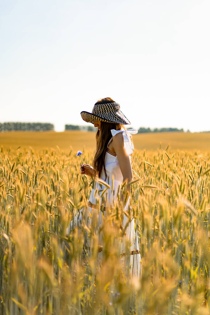 白いドレスに長い髪の若い美しい女性とライ麦と小麦の黄色のフィールドの間で帽子. - 写真・画像