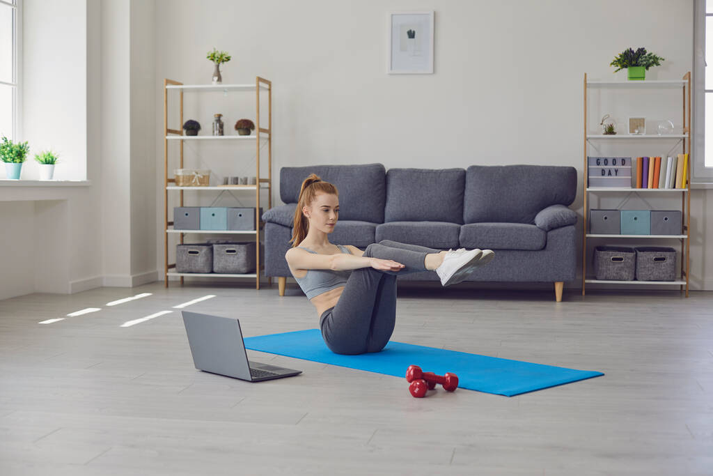 Γυμναστήριο κορίτσι κάνει ασκήσεις στο πάτωμα και βλέποντας ένα βίντεο κατάρτισης, ενώ έχοντας ένα φορητό υπολογιστή σε ένα σαλόνι. Online εκπαιδευτικό βίντεο στο σπίτι. - Φωτογραφία, εικόνα
