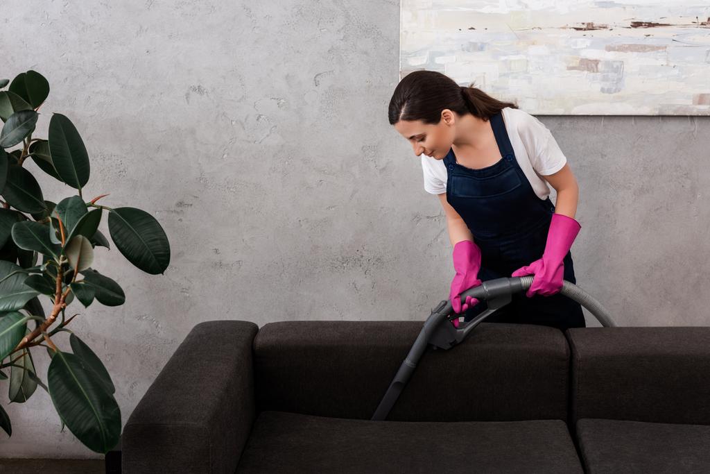 Attraktiver Reiniger in Uniform und Gummihandschuhe reinigen Polster der Couch mit Staubsauger  - Foto, Bild