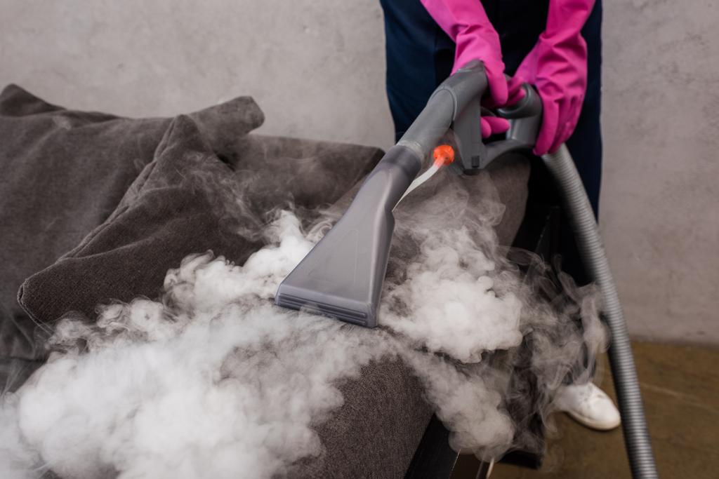Ausgeschnittene Ansicht von Reiniger in Gummihandschuhen mit Staubsauger mit heißem Dampf auf der Couch  - Foto, Bild