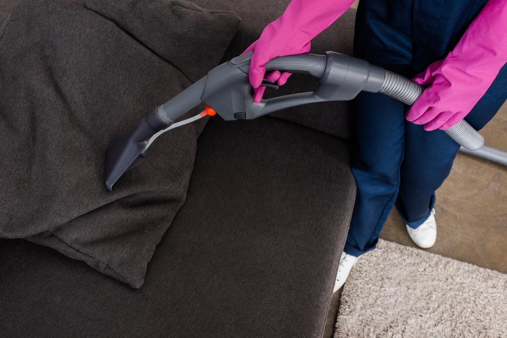 Обрезанный вид чистящего средства в резиновых перчатках с помощью пылесоса на подушке на диване
  - Фото, изображение
