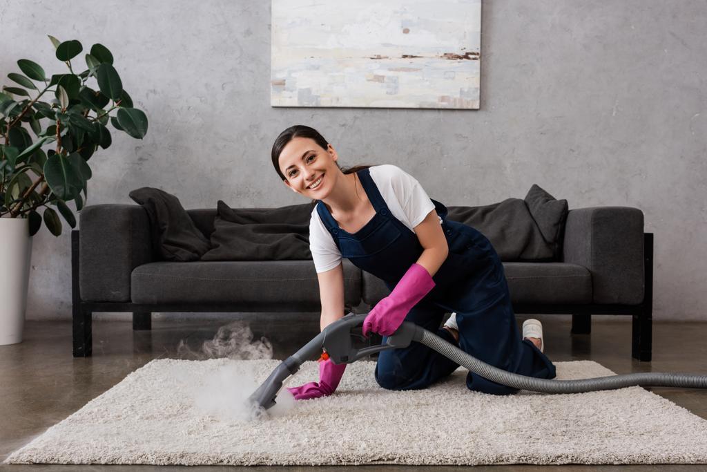 Cleaner sorridente alla fotocamera durante l'utilizzo di aspirapolvere con vapore caldo sul tappeto in soggiorno - Foto, immagini