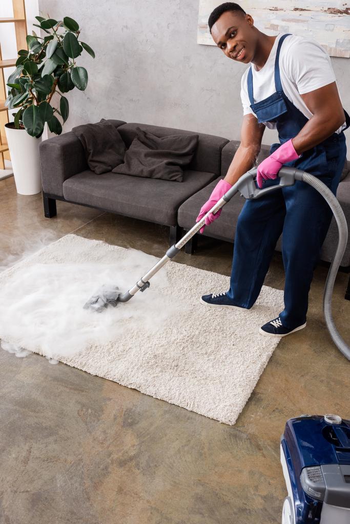 African american cleaner in uniform and rubber gloves smiling at camera while cleaning carpet with hot steam - Photo, Image