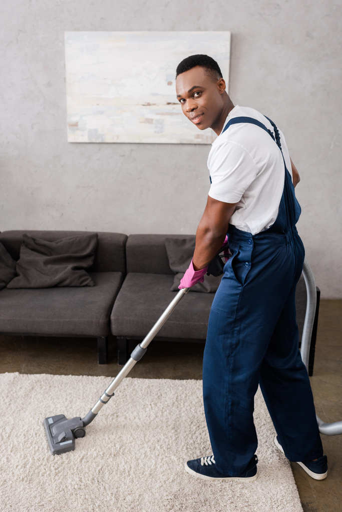 Afrykański amerykański sprzątaczka w mundurze czyszczenia dywanu i patrząc na aparat fotograficzny w salonie - Zdjęcie, obraz