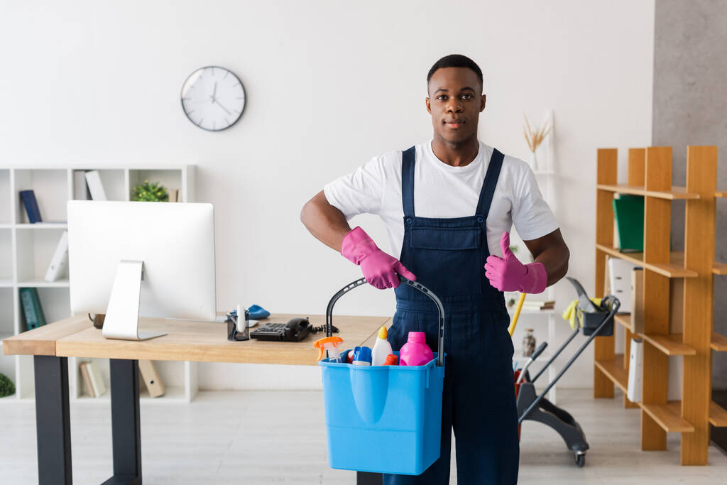 African american cleaner showing thumb up and holding bucket of cleaning supplies in office  - Photo, Image