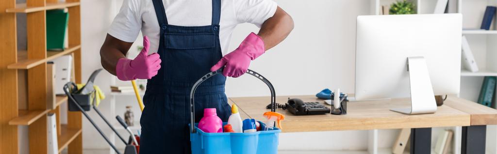 Coltivazione panoramica di detergente afroamericano in uniforme che mostra come e tiene in mano i prodotti per la pulizia in secchio in ufficio - Foto, immagini