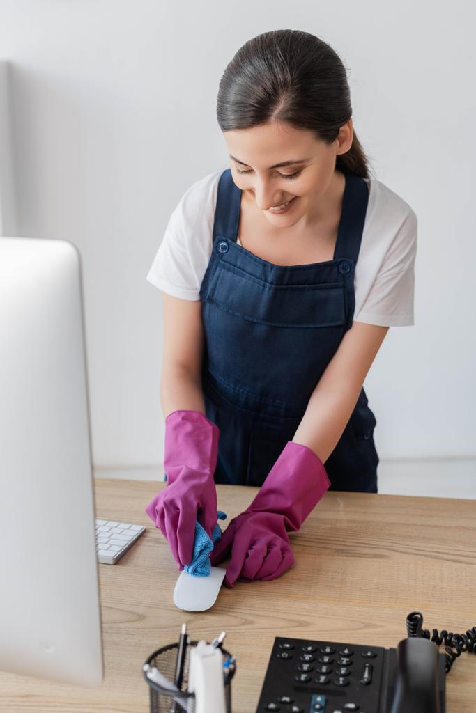 Selective focus of smiling cleaner holding rag while cleaning computer mouse on office table  - Photo, Image